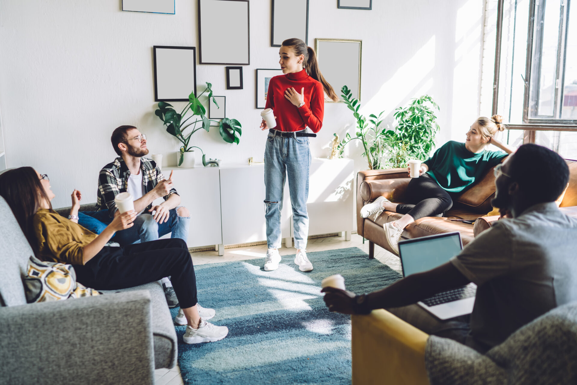 Excited young woman standing with plastic cup of coffee in hand and having conversation with laughing friends sitting around with drinks and gadgets